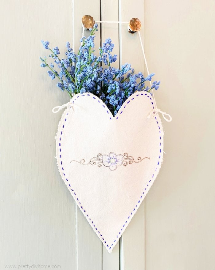 A home decor Valentine craft idea in cream fabric with dark blue embroidery and flowers. A little heart shaped Valentine hanger with lavendar flowers.