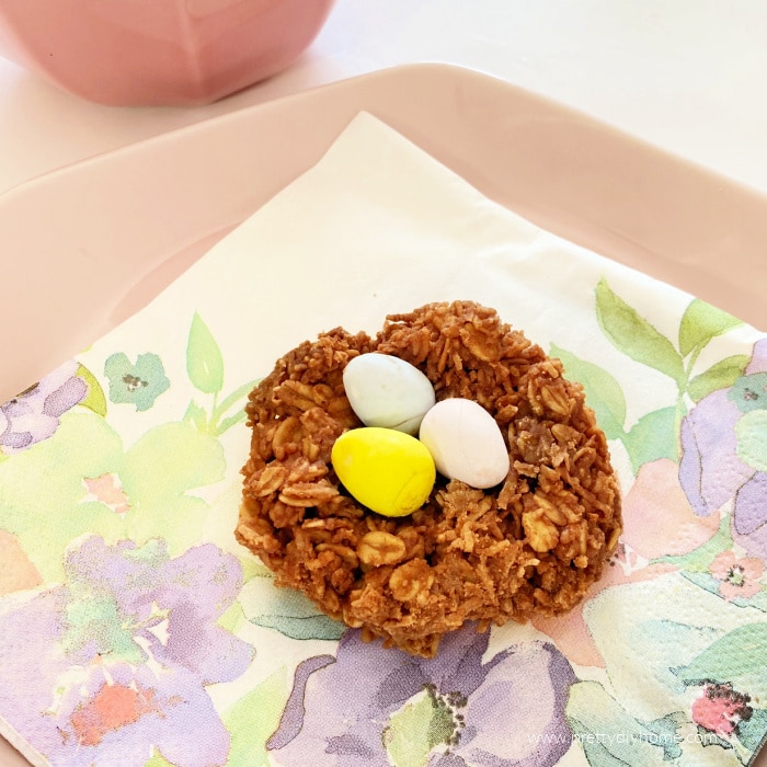 A single Easter cookie made with chocolate and coconut base. The Easter treat is then topped with three mini chocolate Easter eggs so that it looks like a birds nest. The cookie is sitting on a pink plate covered with a pretty floral Easter napkin.