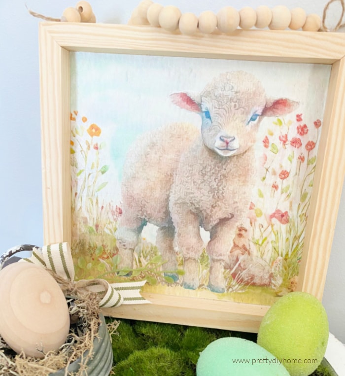 A Spring sign idea with a watercolour lamb, farmhouse bead hanger. It is in soft Spring pastel colours and is sitting on a tray with Easter eggs.