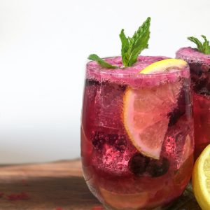 A tall glass filled with a blueberry lemonade recipe. The lemonade is garnished with mint, ice and lemon slices.