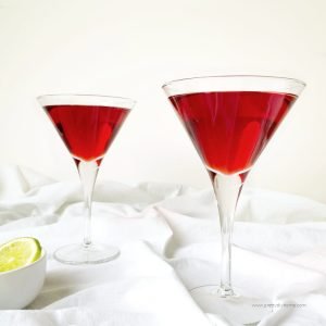 Two cranberry cosmos being served in a martini glass. The cranberry cosmos are a deep red colour and there is a white bowl of lime slices in the background.