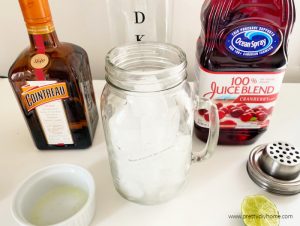 The ingredients for a Cranberry Cosmo Cocktail, including a shaker with ice, real cranberry juice, fresh squeezed lime juice, vodka and Cointreau.