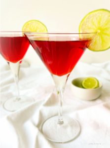 Two Cranberry cosmo cocktails served in martini glasses. They cranberry cosmos are garnished with slices of lime and there is a small bowl of lime slices in the background.