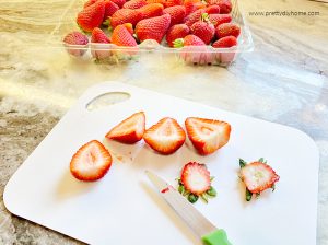 Hulling fresh strawberries after washing. The strawberries are on a white cutting board with a knife and there is a basket of strawberries in the background,