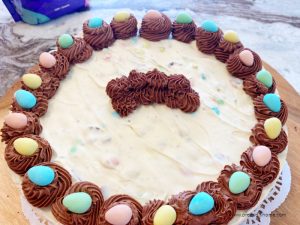 A partially decorated Easter dessert ice cream cake. The image shows the outside swirls of chocolate icing each holding a pretty candy egg. The center shows several swirls of chocolate that will hold the Easter bunny center.