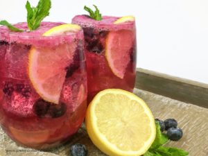 Two glasses filled with ice, lemon slice, crushed mint and blueberry lemonade.