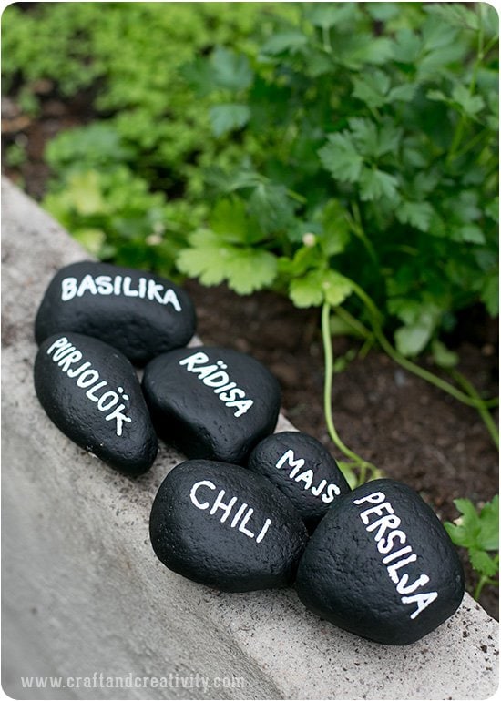 Various sized rocks painted dark black with white plant names lettered on them. They garden markers are sitting on the curb of a garden.