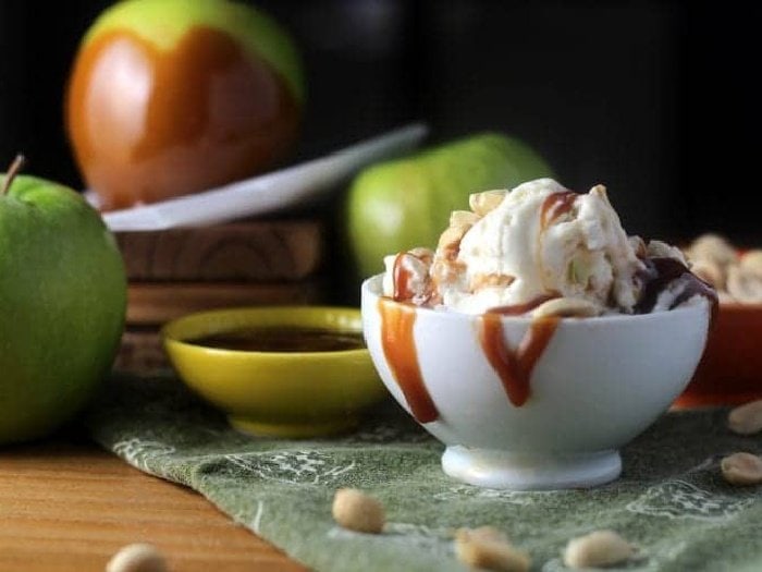 A white bowl filled with caramel apple ice cream.  The ice cream is drizzled with caramel sauce and you can see caramel apples in the background.