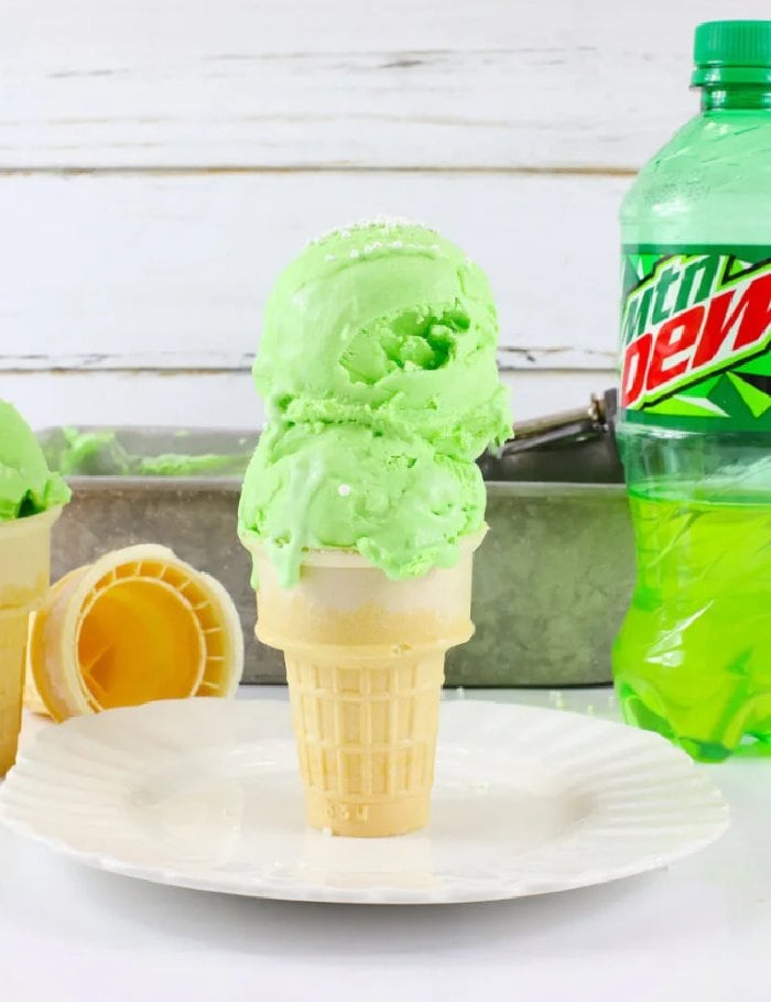 A large double scoop cone of bright green mountain dew ice cream.   There is  a bottle of mountain dew in the background and a white tile backsplash.