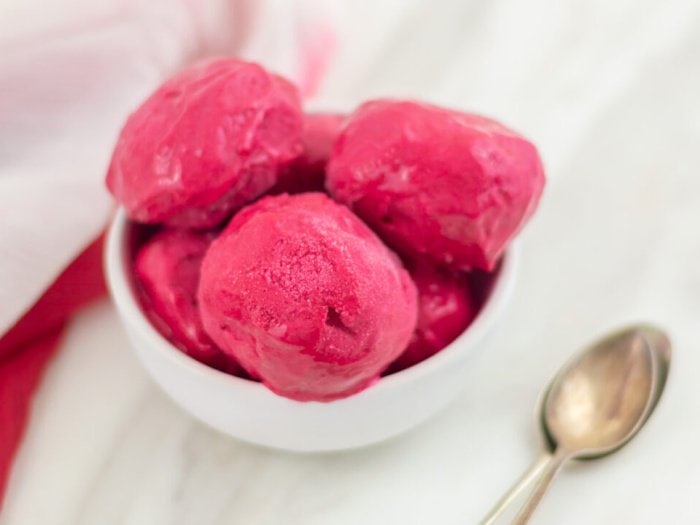 Six small scoops of homemade raspberry ice cream in a white dessert bowl. There is a red napkin, white tablecloth and spoon in the background.