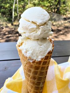Homemade vanilla ice cream being served outside on a hot day so the ice cream is melting a little bit. The ice cream cone has two scoops of homemade vanilla ice cream and is a waffle cone. The background shows the top of a picnic table and green trees.