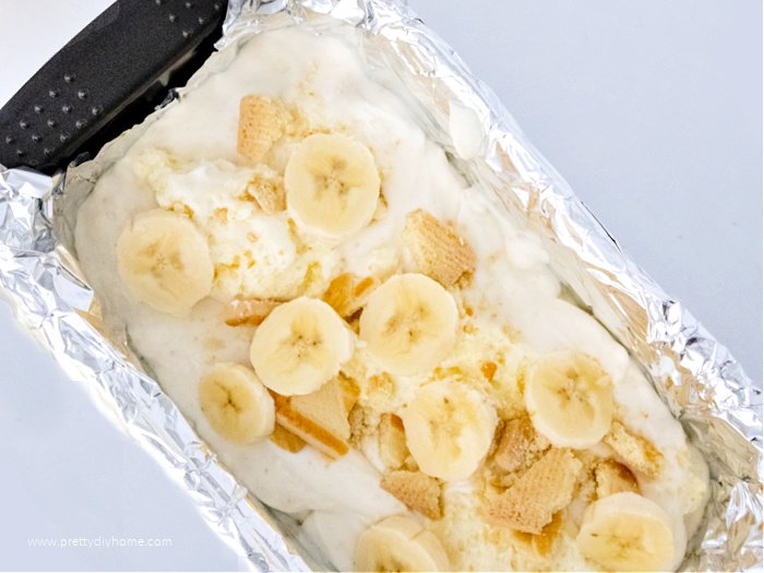 A tin foil lined loaf pan willed with homemade ice cream. The ice cream is bein topped with slice bananas in a layer.