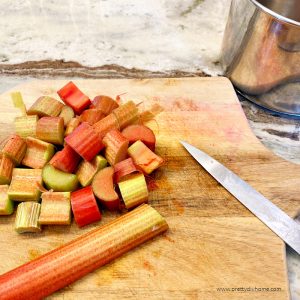 A cutting board covered with fresh rhubarb. They rhubarb stalk is being cut into small chunks prior to cooking.