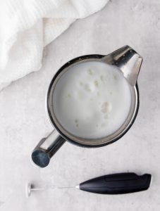 A barista mug and a frothier. The mug is filled of warm frothed milk for making a vanilla latte at home.