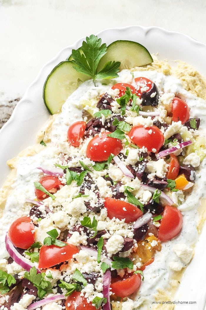 Layered Greek dip with hummus, tzatziki, feta, red onions, cherry tomatoes served on a white platter with a granite counter backdrop.