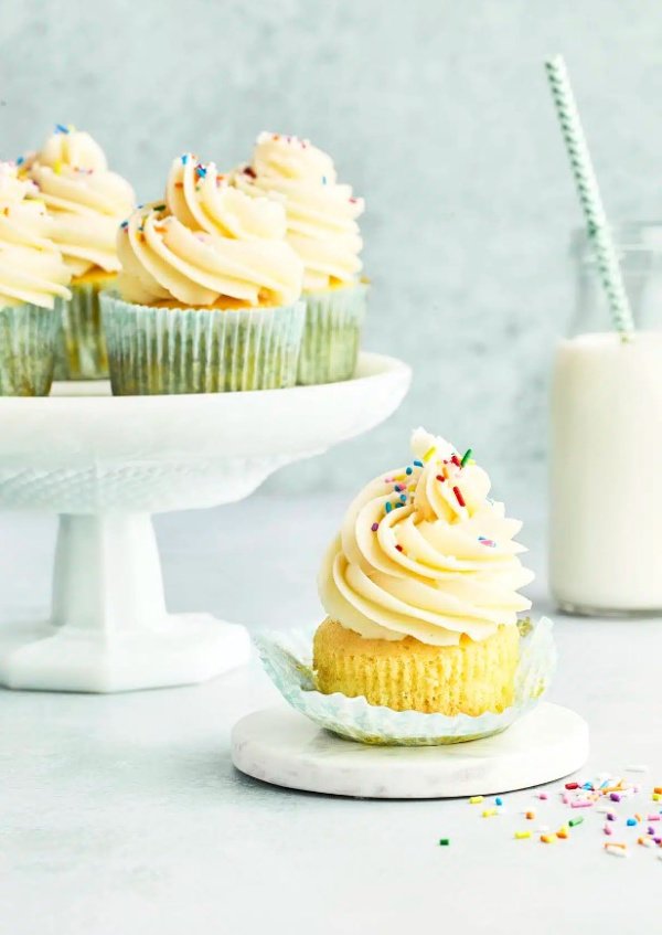 A white pedestal plate with four vanilla cupcakes covered in large swirls of American buttercream and sprinkles. There is a single cupcake being served and a milk glass with a straw in the background.