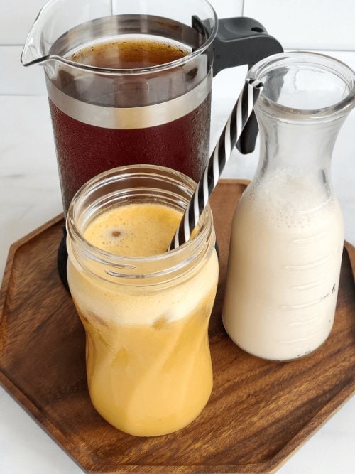 A coffee press, a glass jar of milk and a pumpkin cold spiced coffee are sitting on a brown wood tray. The pumpkin cold coffee has a straw in it.