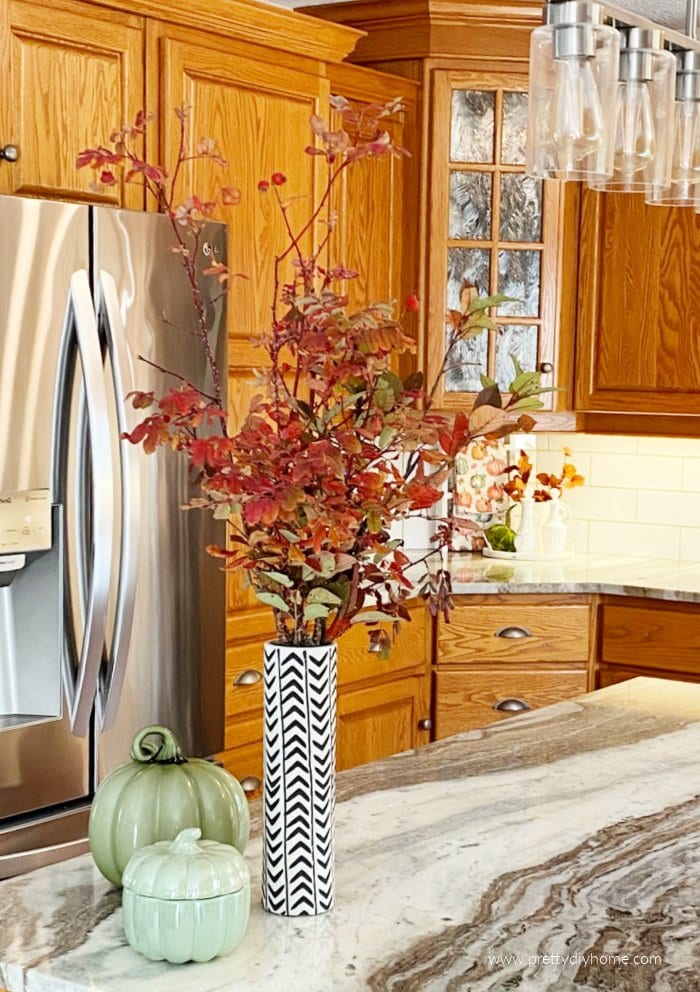 An inexpensive Fall decor idea with a large bouquet of live Fall branches in a vase. The vase of orange leaved branches is sitting on the island of a kitchen.