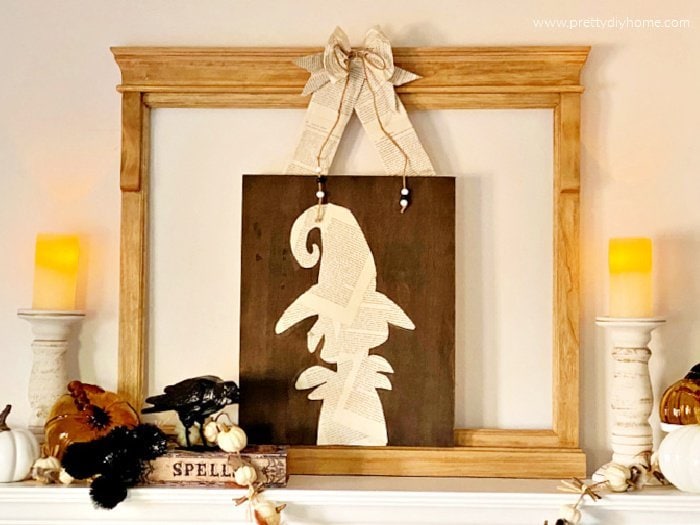 A witchy halloween mantel decorated with a diy halloween witch made with dollar store frame and book pages. The witchy halloween decoration is hanging from a large wood frame and is surrounded by candles, pumpkins a spell book and a lovely black crow.