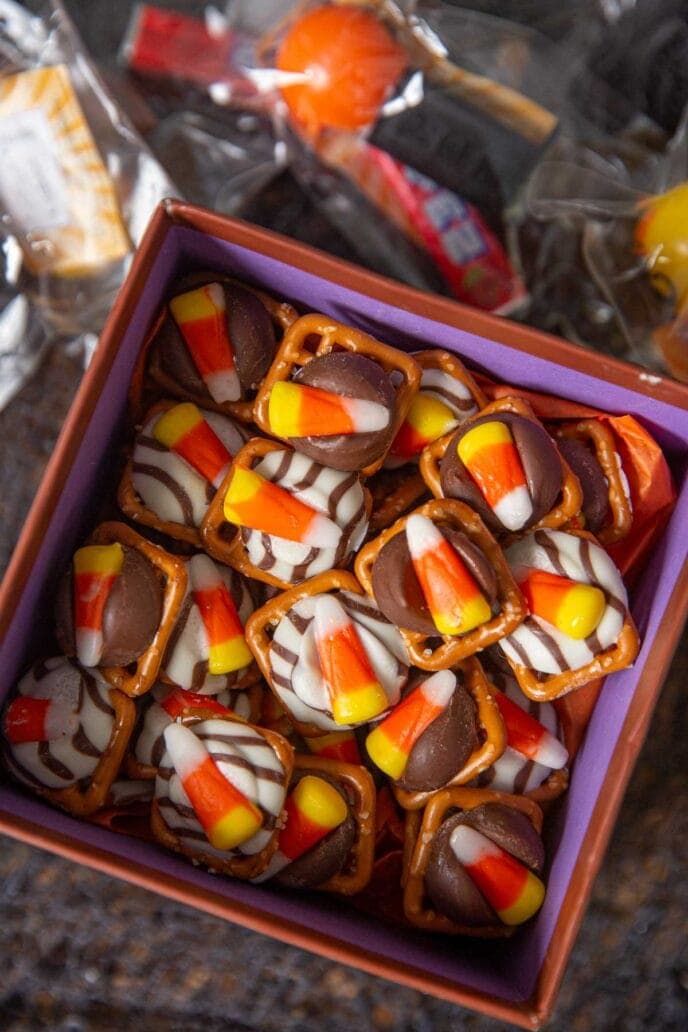 A small orange and purple box filled with halloween pretzel hug candy.  Each halloween treat is made up of a square pretzel with a hey melted on top and then a candy corn.