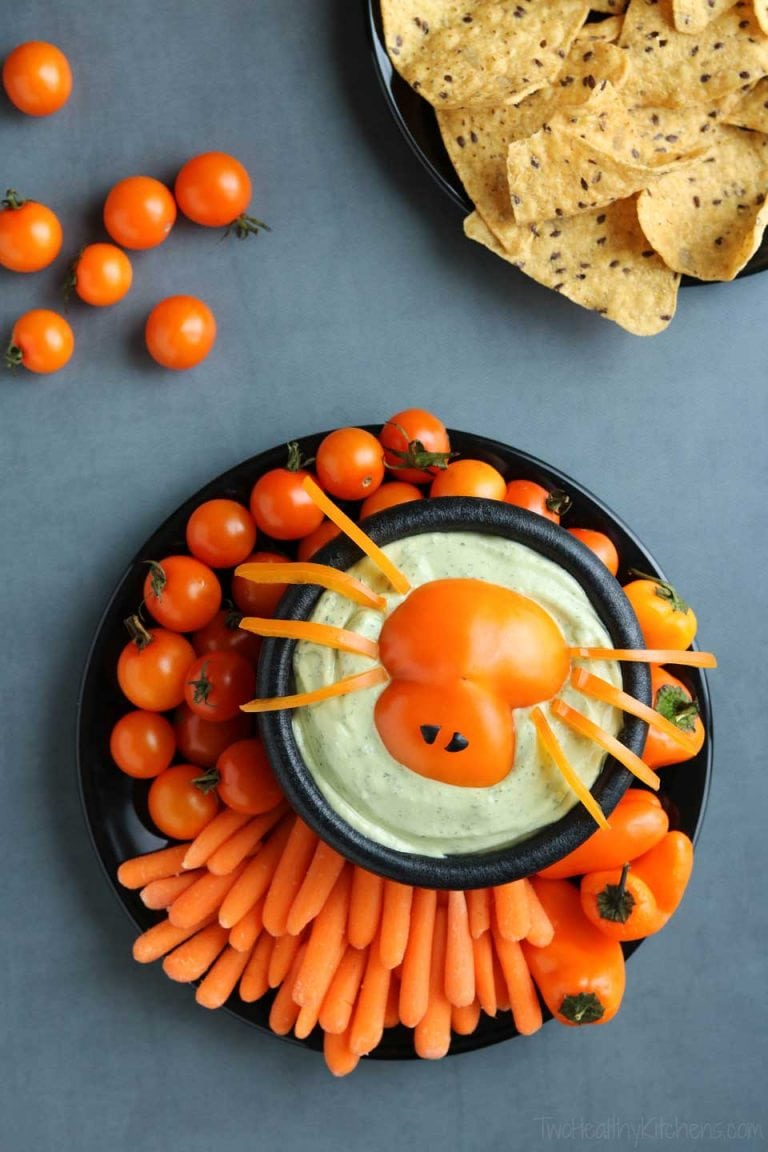 A vegetable tray served on a black tray and bowl.  This Halloween appetizer has a bowl of green dip with a cutout tomato onto that looks like the body of a spider, and the legs are thin cuts of orange peppers.