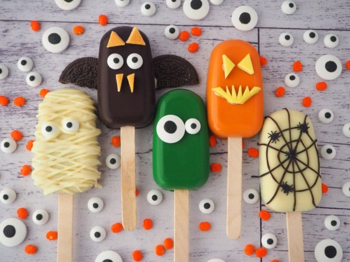 Five different halloween cakesicles. There is a mummy, a bat, a green monster an orange pumpkin and a spider web cakesicle.