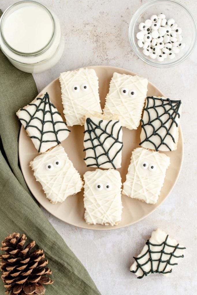 A round cream plate covered with Halloween rice krispie treats.  There are six rice krispie treats that look like mummies and four that have black spider webs on them.  