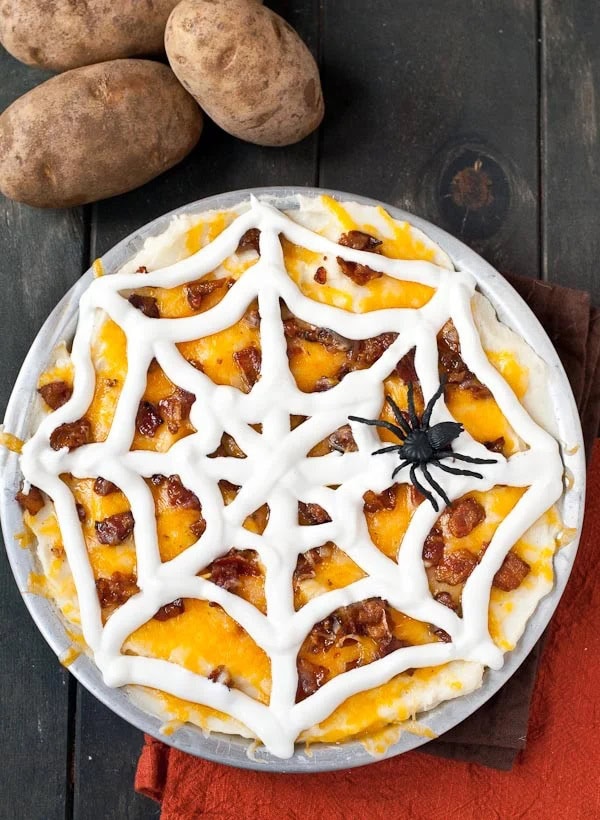 A loaded mashed potato casserole topped with cheese, then a sour cream spiderweb, then decorated with a large plastic spider.