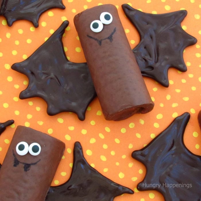 Chocolate Halloween bats with snack  cake bodies, candy eyes, chocolate fangs, and solid chocolate wings.  There are four bats laying against a orange polka dot background.