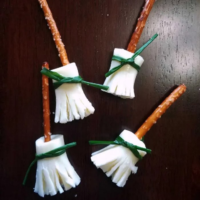 Four witch broom appetizers with pretzel broom stick handles sliced cheese bristles tied with green chives.