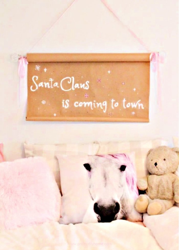 A brown paper scroll sign for a kid's bedroom The Christmas scroll sign is hanging above the bed as Christmas decor in a little girl's bedroom. The Christmas sign diy says Santa Claus is coming to town. The remaining decorations include a pink furry pillow, pink horse pillow and a vintage teddy bear sitting on a bed covered with a white fur bedspread.