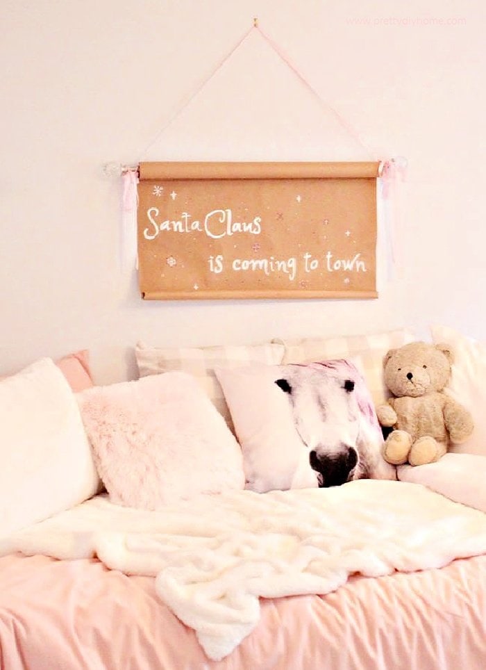 A Christmas sign diy craft hanging over a little girl's bed for Christmas. The Christmas scroll is made using brown paper and dollar tree supplies. It is sitting over the head of a little girls bed thats covered with pink furry cushions, a teddy bear and pink bedding.