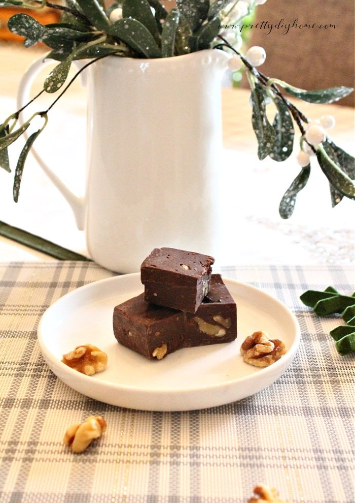 Three pieces of chocolate fudge recipe sitting on a white plate surrounded by shelled walnut halves and Christmas decorations in white and green. There is a white pitcher in the background with a velvet christmas ornament in the background.