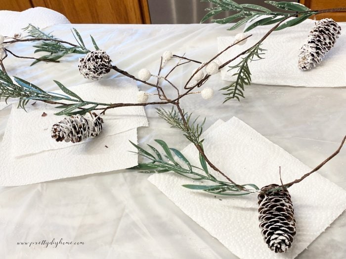 A dollar store garland with a few green leaves, white sparkling beads, and pinecones. The garland is lying on the counter while the pinecones are being painted white.