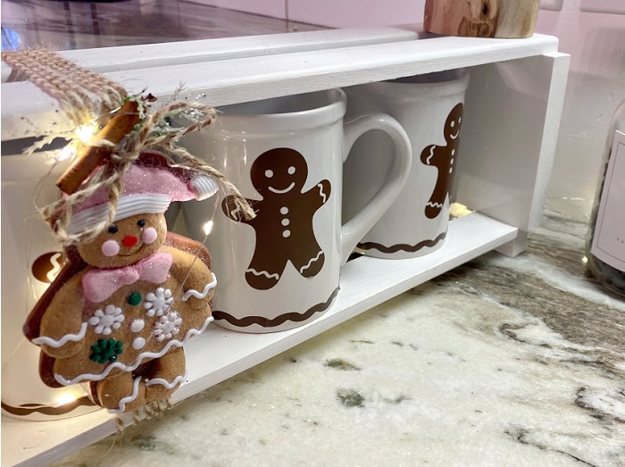 Gingerbread Christmas coffee and hot cocoa bar with white mugs, wooden caddy, fairy lights, and labeled jar.
