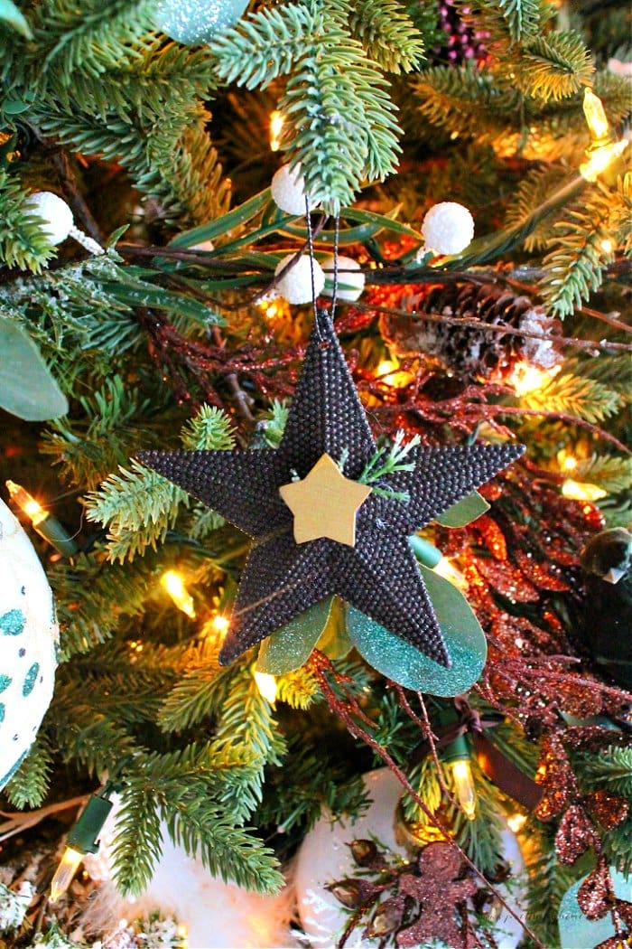 A striking and quaint dollar tree christmas ornament in a star shape. The star is painted a dark chocolate brown and features a metallic gold miniature star in its center. The Christmas tree ornament is hanging on the tree surrounded by other neutral Christmas decor, in browns, greens and whites.