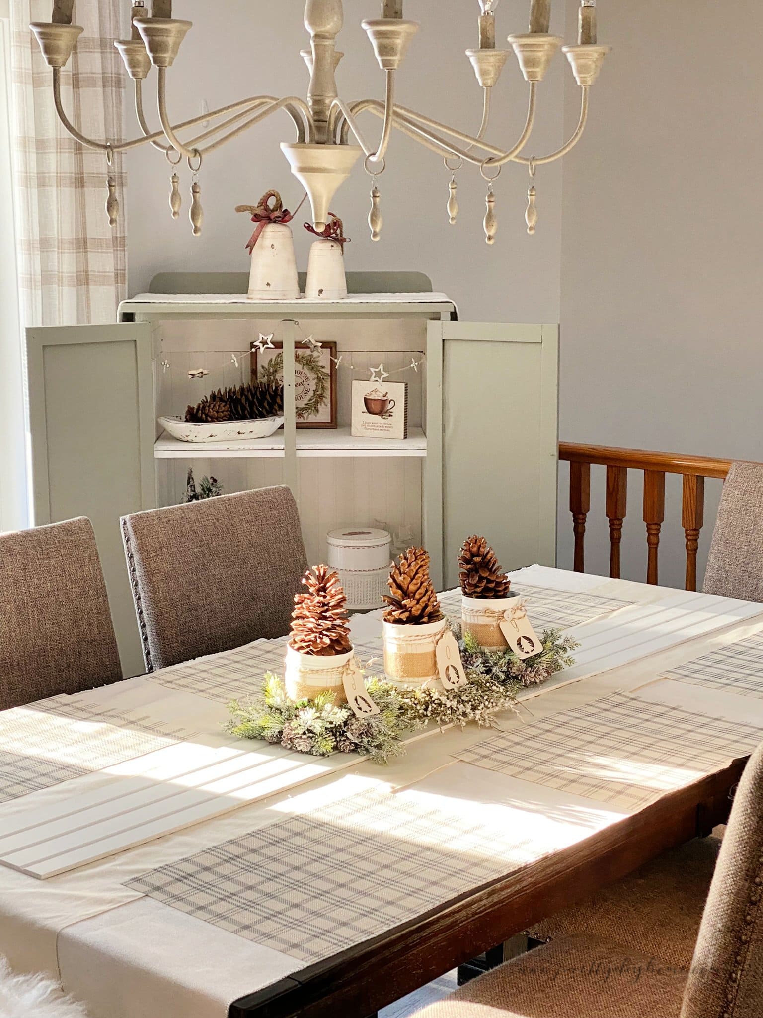 A dining room decorated for Christmas in soft neutral colours like cream, sage and white. The feature is a beautiful tin can craft Christmas centerpiece with pinecones and greenery.