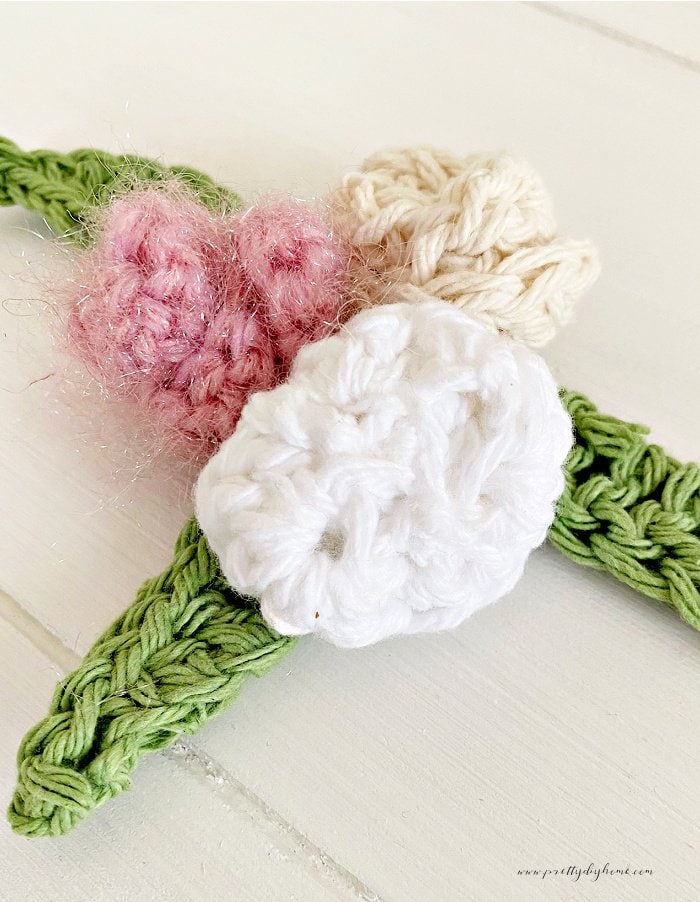 Three tiny crochet roses.  One is pink, one is white and one is pink  They are accessorized with spring green coloured crocheted leave.  The background is white wood.