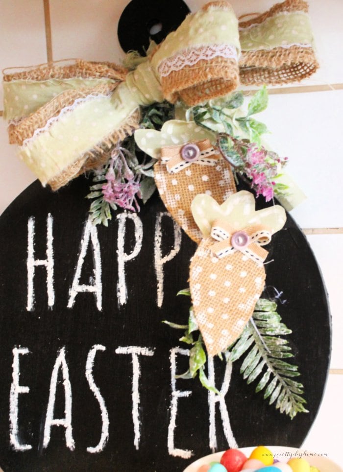 A DIY Easter chalkboard with bright white lettering that says Happy Easter. A lovely burlap bow, and fabric carrots.