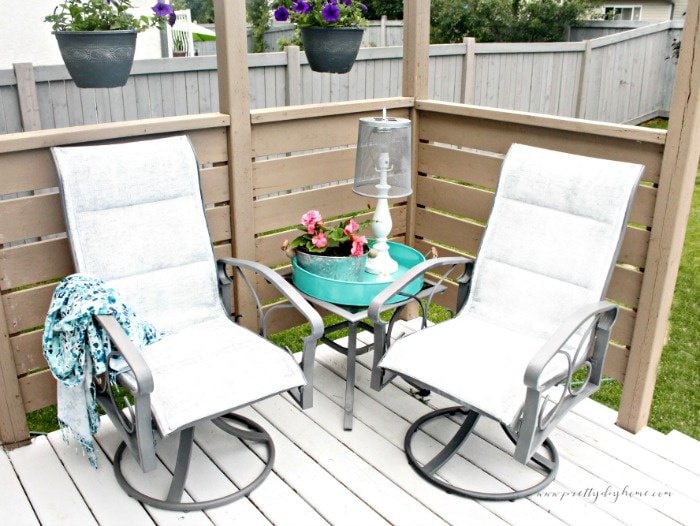 A white DIY outdoor solar lamp with a metal shade sitting on an end table. The lanter has a white patio chair on each side.