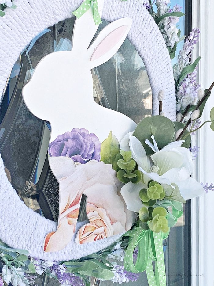 A closeup of a DIY Spring wreath Dollar Tree craft with a large white wooden bunny, floral motive and a large wreath with white rope, purple dollar tree flowers and a beautiful magnolia flower.