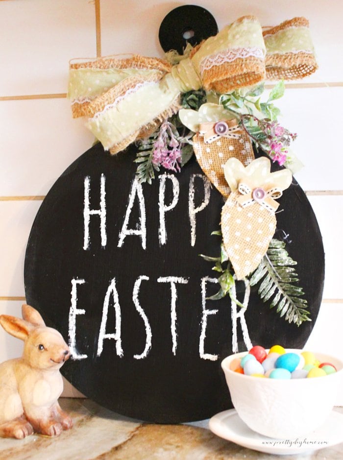 A dollar store DIY Easter chalkboard that says Happy Easter.  It is sitting on a kitchen counter with a large burlap bow, and fabric carrots embellishments.