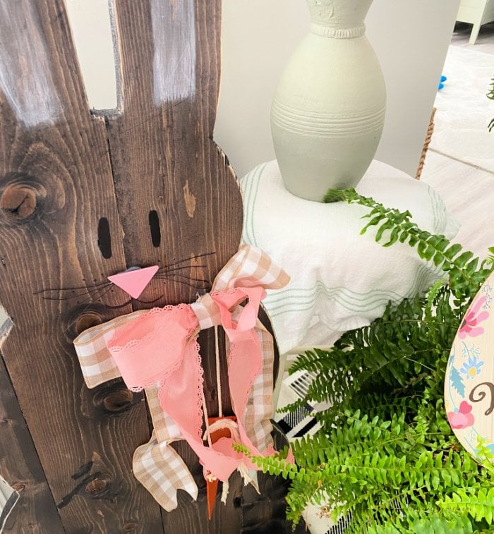 A close up of a wooden bunny face showing eyes painted with a black sharpie, a pink painted triangular nose made of wood, and thin black wire whiskers.