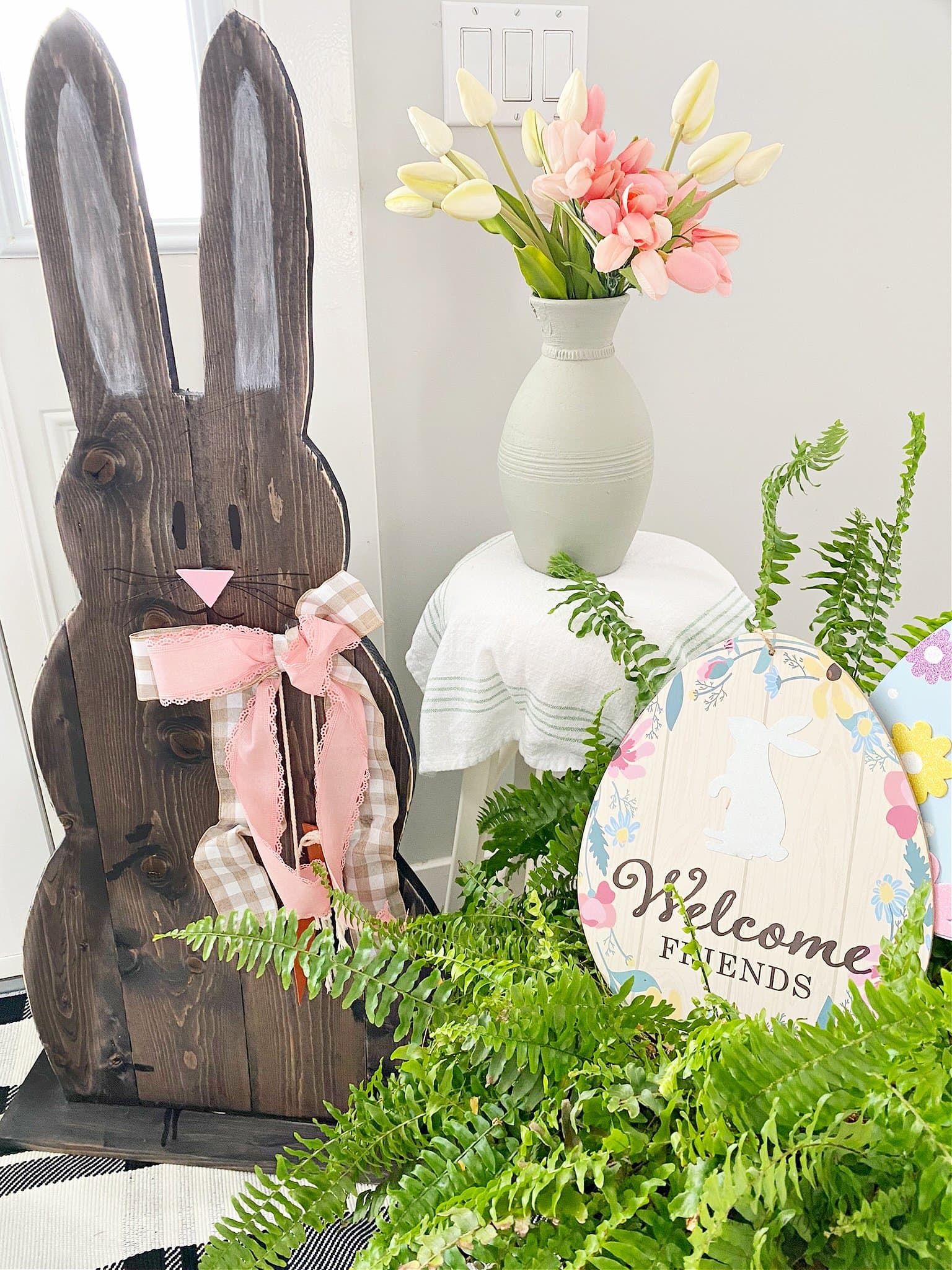 A homemade wooden bunny DIY in chocolate brown with a pink bow, sitting beside a large planted fern with Easter eggs and a jar of tulips.