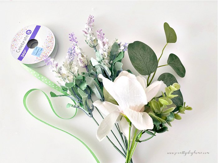 Faux magnolia flower with green leaves, a thin green polka dot spool of ribbon and some small purple Spring flower sitting on a white background before being added to a Spring wreath craft for the front door.