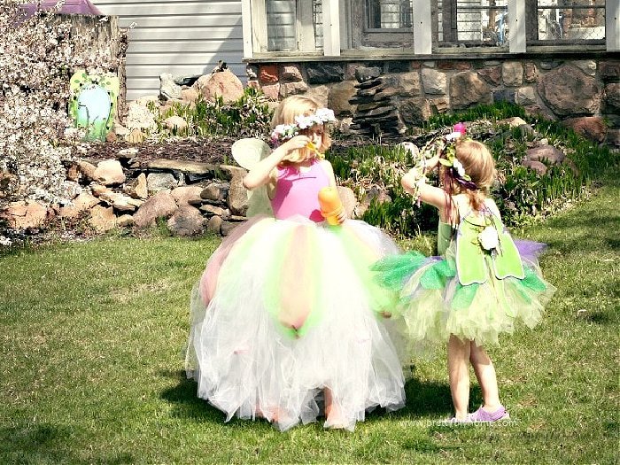 Two little girls playing in no sew tutus for kids, one is pink and white the other is greens and purples. Both little girls also have wings and flower crowns.