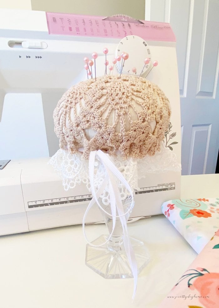 A DIY pin cushion with crocheted top, and a lace bottom. Its holding a dozen pink sewing pins and is sitting in front of a sewing machine. The pin cushion has a crystal stand.