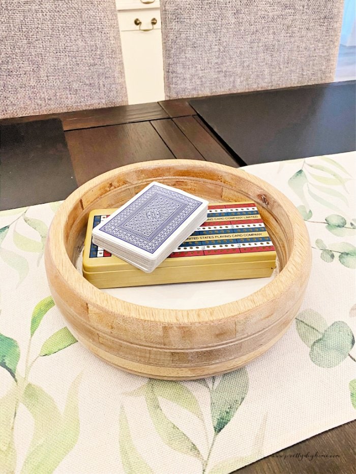 A vintage wood bowl in light birch with a painted white bottom. The wooden bowl is holding a crib board and cards and is sitting on a white and green table runner.