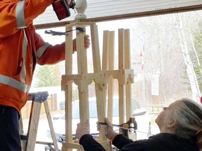 Two people placing a top onto an obelisk.  One is standing on a ladder with a drill, the other is handing clamps.