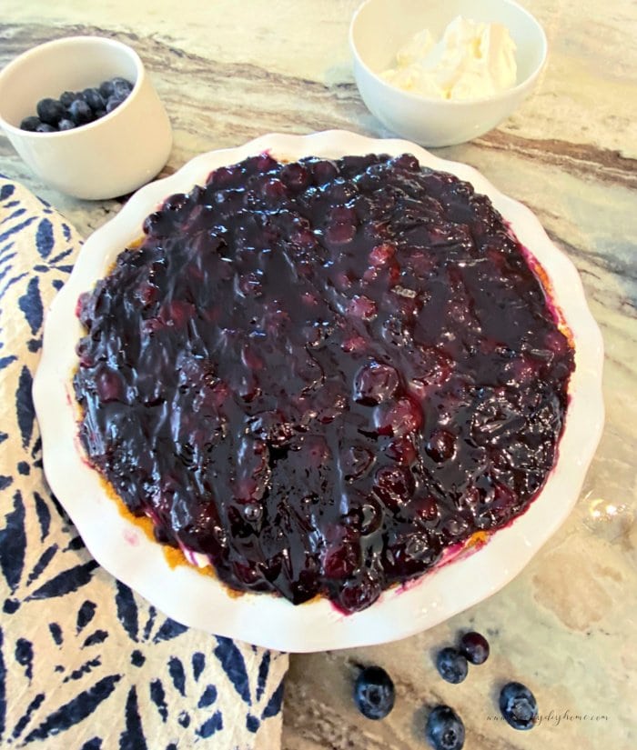 A large no bake blueberry cream cheese pie sitting on a counter in a white porcelain pie plate. The top has a layer of dark purple blueberry topping and you can see the graham cracker crust around the outside.